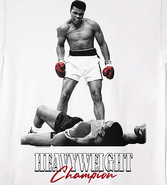 A white short-sleeve crewneck t-shirt featuring a graphic print of boxer Muhammad Ali standing over a defeated opponent, with the text 'HEAVYWEIGHT Champion' printed below in black and red.