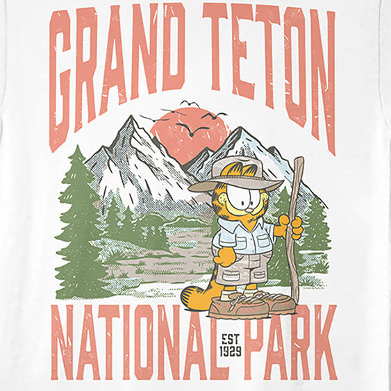 A white short-sleeve crewneck t-shirt featuring a colorful graphic print of Garfield hiking in front of mountains with the text 'GRAND TETON NATIONAL PARK' in coral pink.