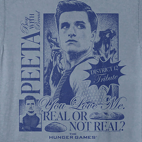 A light blue crewneck t-shirt featuring a dark blue graphic print of Peeta from Hunger Games, with text including 'PEETA', 'You Love Me. REAL OR NOT REAL?', and 'District 12 Tribute'.