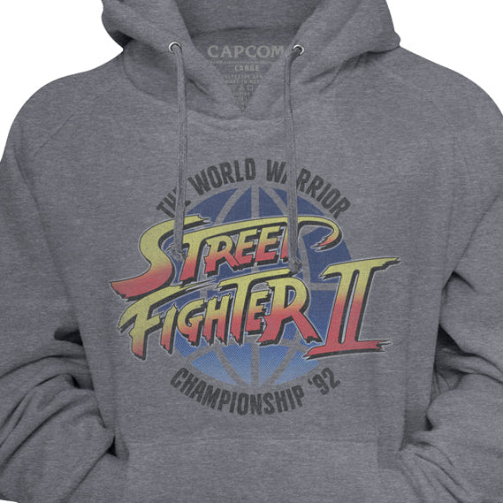 A heather gray pullover hoodie featuring the colorful 'Street Fighter II' World Warrior 'B' Championship '92' graphic on the chest, with a visible Capcom logo printed inside the collar.